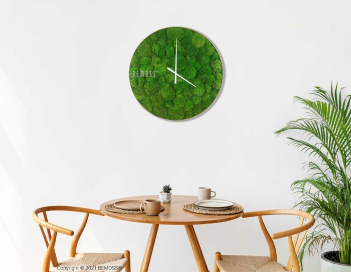 Une horloge murale ronde avec un cadran recouvert de mousse et des aiguilles blanches est montée sur un mur blanc. En dessous, une petite table en bois avec deux chaises contient un ensemble minimaliste composé d'une petite plante en pot, d'une tasse et d'une assiette. Une plante verte trône à côté de l’Horloge de mousse BOLMOSS Light sur le côté droit de la scène.