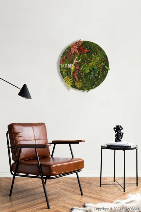 Un salon moderne comprend un fauteuil en cuir marron à côté d'une table d'appoint noire contenant une petite sculpture. Sur le mur de couleur claire, il y a un Tableau de mousse rond PLANTES Loreto avec de la mousse et des feuilles, et une lampe angulaire noire est positionnée à gauche.