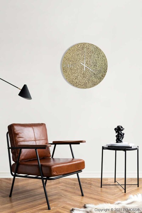 Un espace de vie moderne comprenant un fauteuil en cuir marron avec une structure en métal, une petite table d'appoint noire ornée d'une sculpture et une Horloge de mousse en mousse de RENNE unique. La zone abrite des murs végétaux, conçus avec une esthétique minimaliste et comprend une élégante lampe anglepoise noire.