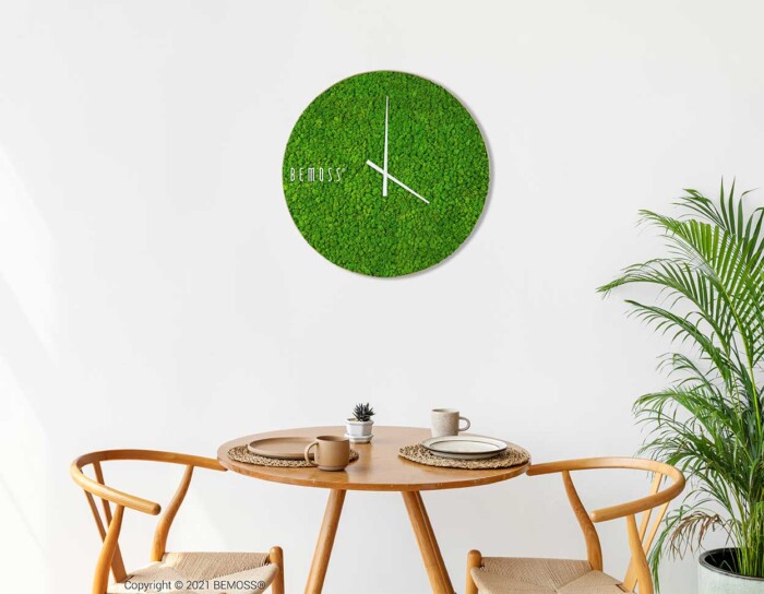 Un coin repas moderne comprend une table en bois et deux chaises. Au mur est accrochée une Horloge de mousse en mousse ronde de RENNE avec des aiguilles blanches et un design minimaliste. Une plante en pot est placée sur la table avec deux assiettes, et une plante se trouve à droite de la table.