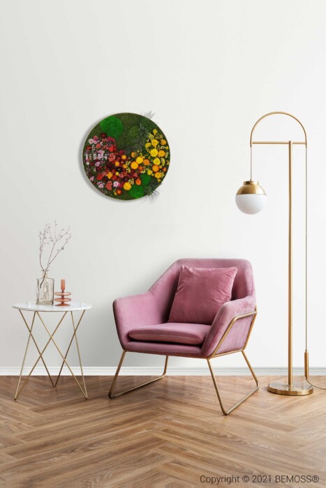 Un salon moderne comprend une chaise rembourrée rose avec des pieds dorés, un lampadaire doré avec un abat-jour sphérique et une petite table d'appoint blanche contenant des objets décoratifs. Une œuvre d'art florale circulaire est montée sur le mur au-dessus de la chaise, rehaussée par un vibrant Tableau de mousse rond PLANTES Parra.