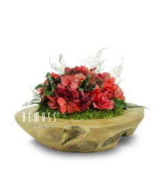 Un arrangement floral de fleurs rouges et de verdure est présenté dans un bol en bois rustique de forme ovale. La texture et les courbes naturelles du bol complètent les couleurs vibrantes des fleurs. Les mots "Bol en teck Celada Ø30cm" sont imprimés sur le côté gauche du bol, ajoutant une touche unique inspirée des Murs végétaux.