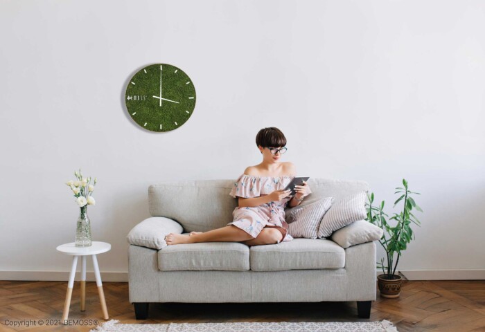 Une femme vêtue d'une robe à fleurs est assise sur un canapé beige en train de lire un livre dans un salon minimaliste. Une Horloge de mousse en mousse de RENNE verte avec cadran orne le mur blanc, accompagnée d'une petite table d'appoint blanche avec un vase en verre et une plante en pot au sol.