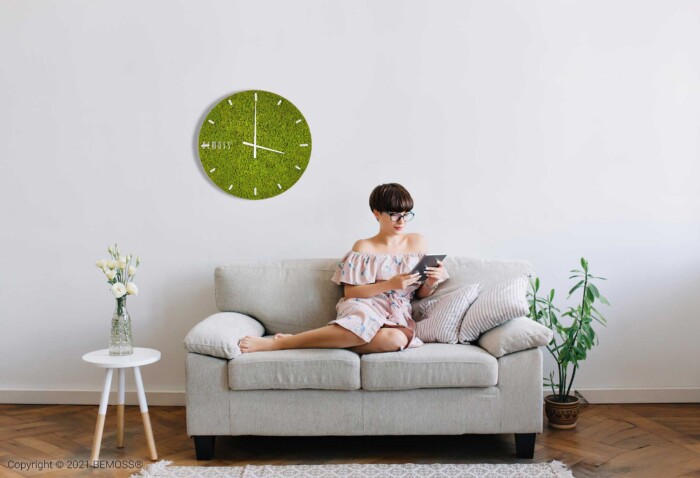 Une personne est assise sur un canapé gris clair dans un salon minimaliste et lit un livre. Le mur derrière eux présente une grande Horloge de mousse en mousse de RENNE verte avec cadran. À gauche se trouve une petite table avec un vase de fleurs et à droite une plante en pot. Le décor évoque l'ambiance d'élégants murs végétaux.