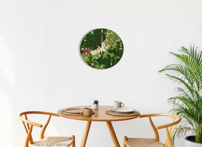 Une salle à manger minimaliste comprend une Horloge de mousse BEMOSS® ORTHO SPRING avec cadran, deux chaises en bois avec sièges tressés, une table ronde en bois contenant un petit vase gris, une plante en pot et un cahier. À droite de la table se dresse une grande plante verte devant des murs en mousse vibrants, ajoutant de la texture à l'espace.