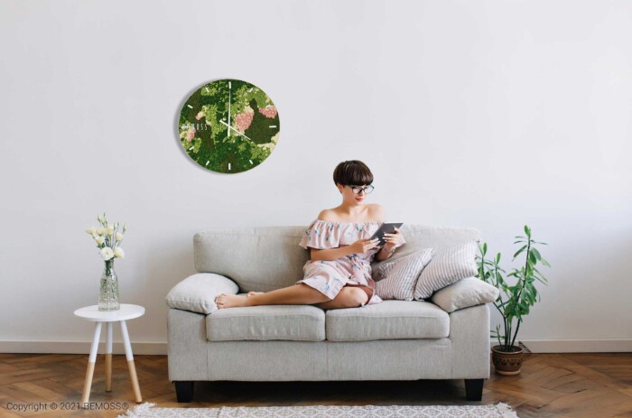 Une femme portant des lunettes et une robe à fleurs rose est assise sur un canapé gris clair tout en lisant une tablette. La pièce minimaliste présente des murs blancs ornés d'une Horloge de mousse BEMOSS® ORTHO SPRING avec cadran et de murs végétaux vibrants. Une petite table blanche avec un vase de fleurs et des plantes en pot se trouve à proximité.