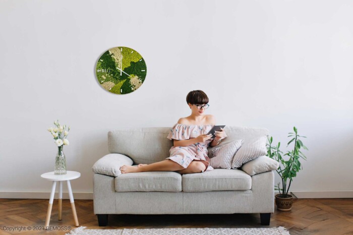 Une femme est assise sur un canapé gris clair dans un salon minimaliste. Elle porte une robe à épaules dénudées et lit un livre. Une Horloge de mousse BEMOSS® ORTHO SPRING avec cadran est suspendue au-dessus d'elle, complétée par des murs végétaux et des plantes d'intérieur. A proximité, il y a une petite table d'appoint ronde avec un vase.