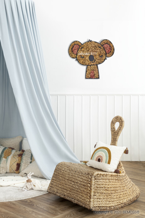 Une chambre d'enfant confortable comprend une chaise longue en osier ornée d'un coussin arc-en-ciel, placée à côté d'un rideau bleu clair. Un tableau de mousse ressemblant à un koala est suspendu au-dessus des lambris blancs, ajoutant une touche fantaisiste à l'espace.