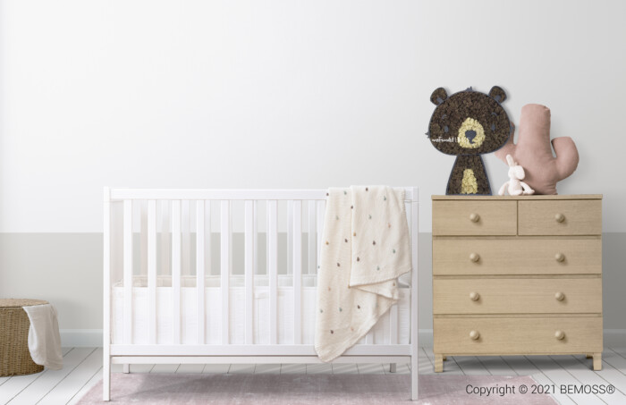 Une chambre de bébé minimaliste comprend un berceau blanc avec une couverture à motifs beige drapée sur la barrière. A côté, une commode en bois clair accueille un gros ours en peluche marron et un lapin en peluche rose. Un panier en osier repose sur le sol à proximité, tandis que des murs végétaux ajoutent une touche de verdure vibrante. La pièce est doucement éclairée.