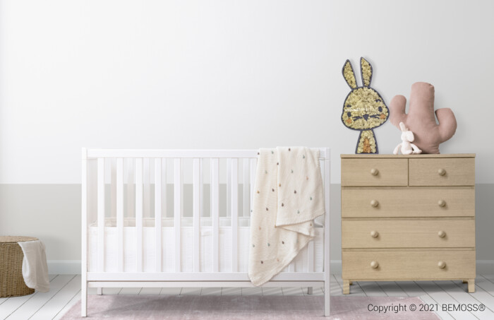 Une chambre de bébé minimaliste comprend un berceau blanc avec une couverture beige drapée sur le côté. À droite, une commode en bois clair présente un lapin et un cactus décoratifs, avec un petit jouet en peluche à la base. Un panier tressé se trouve sur le sol à gauche du berceau, à côté des murs luxuriants BEMOSS ajoutant une chaleur naturelle.