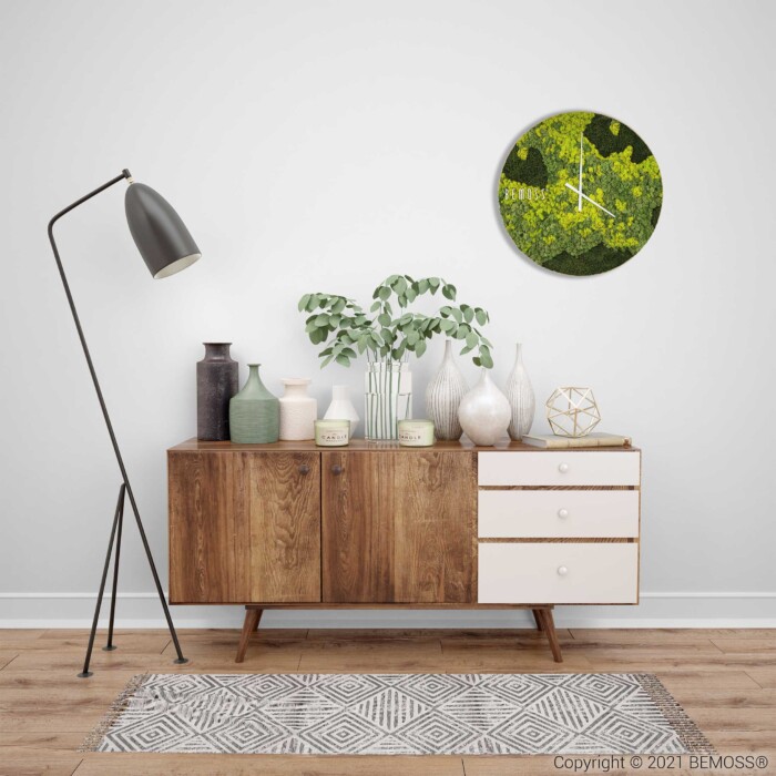 Une scène de salon moderne comprenant un buffet en bois surmonté de divers objets décoratifs, notamment des vases et une petite plante. Un grand lampadaire noir se dresse à gauche. Au-dessus du buffet est suspendue une Horloge de mousse ronde BEMOSS® ORTHO LIME représentant un feuillage vert. Un tapis à motifs repose sur le sol.