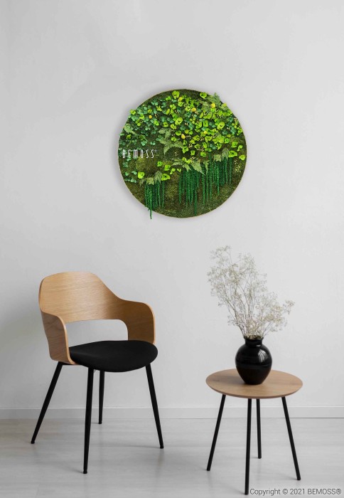 Une chambre moderne et minimaliste comprend une chaise en bois avec un coussin noir à côté d'une petite table ronde contenant un vase noir avec des fleurs blanches. Ci-dessus, une œuvre d'art murale ronde à base de verdure de BEMOSS intitulée 