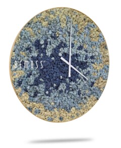 Une horloge murale ronde portant le nom de la marque « BEMOSS » à gauche, présentant un design abstrait composé de motifs texturés bleus, beiges et gris rappelant les murs végétaux. L'horloge a deux fines aiguilles blanches indiquant l'heure. Le fond est blanc, créant un effet d'ombre sous l'horloge.