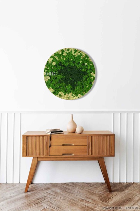 Un buffet moderne en bois aux pieds inclinés se dresse contre un mur blanc aux lambris verticaux. Au-dessus, une œuvre d'art ronde en mousse verte de BEMOSS ajoute une touche de nature. Deux vases beiges minimalistes et un livre reposent sur le buffet, tandis que le sol présente un motif à chevrons.