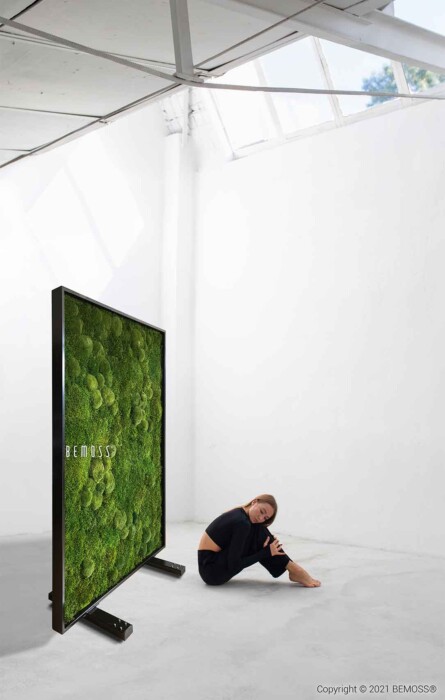 Une femme vêtue de noir est assise sur un sol spacieux et lumineux dans une pièce minimaliste. À côté d'elle se trouve un grand présentoir vertical autoportant avec une surface recouverte de mousse et le mot « BEMOSS », ressemblant à un superbe tableau de mousse. La pièce présente de hauts plafonds et des murs d'un blanc immaculé.