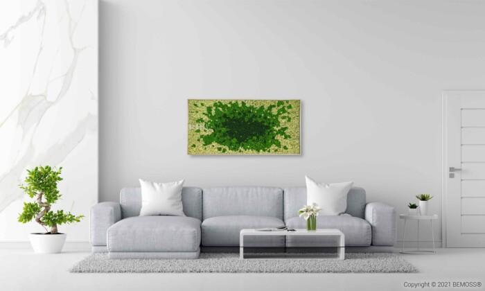 Un salon moderne avec un canapé gris clair et deux oreillers blancs, une table basse blanche avec un vase à fleurs et une plante à gauche. Un tableau de mousse encadré vert est accroché au mur au-dessus du canapé. L'espace est minimaliste et lumineux avec une porte blanche sur la droite.