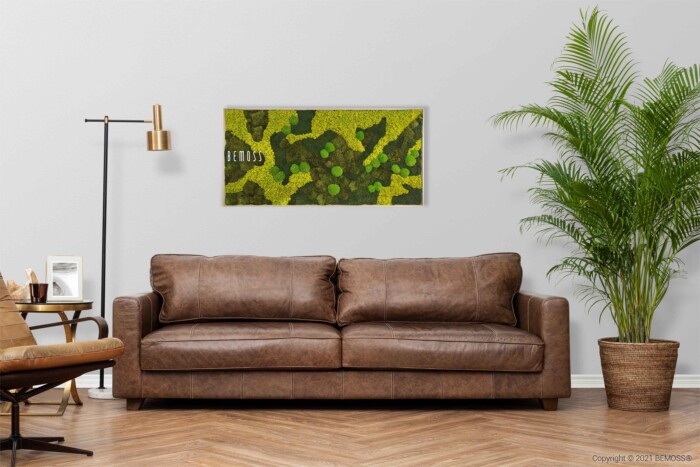 Un salon moderne avec un canapé en cuir marron, une plante en pot à droite et une œuvre d'art murale en mousse verte au-dessus du canapé. Un lampadaire et une petite chaise sont placés sur le côté gauche. L'art mural indique 